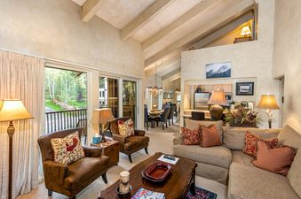 A spacious and inviting living area features warm lighting, comfortable seating, and a dining area, with a balcony offering a view of the natural surroundings.