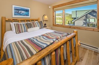 A cozy bedroom in Big Horn 43 features a rustic wooden bed with colorful striped bedding and a large window overlooking a forest and mountains.