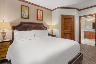 A king-sized bed with a brown leather headboard and white bedding, flanked by two bedside tables with lamps, is featured in the bedroom of Elkhorn Premier 8405, with an en suite bathroom visible through an open doorway.