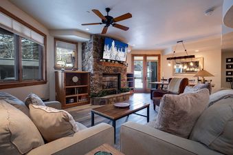 The living room features a stone fireplace, flat-screen TV, comfortable seating, and large windows with views of the outdoors, offering a cozy and inviting space.