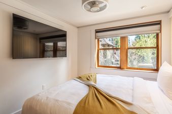 A bright bedroom in Cedar Crest Mallard Cottage 5 with a large window overlooking trees, a wall-mounted TV, and a bed with white linens and a yellow throw.