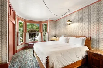 A cozy bedroom with a large bed, patterned wallpaper, a bay window overlooking trees, and an antique pendant lamp.