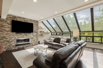 The living area features a comfortable setting with a large screen TV mounted on a stone wall above a modern fireplace, with large windows offering a lush green outdoor view.