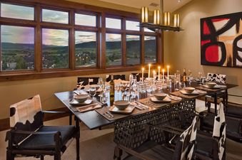The dining area within Flat Tops Peak Penthouse One Steamboat Place 701 features a large dark wood table set with candles and formal place settings, surrounded by unique cowhide chairs, all beneath a modern linear chandelier.