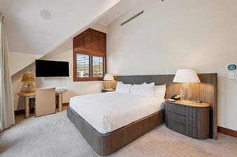 A cozy bedroom in Solaris PHHW features a large bed with a white comforter and pillows, flanked by elegant bedside tables with lamps, a window with wooden shutters, and a television with a desk and chair.