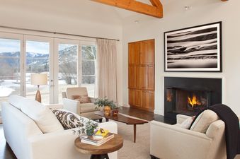 An elegant and cozy living room at Pines Garden 4050 with white sofas, a lit fireplace, and large French doors revealing a snowy mountain landscape.