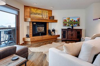 A cozy living room at Ranch at Steamboat RA203 with a fireplace, TV, and balcony access with a BBQ grill, showcasing a snowy mountain view.
