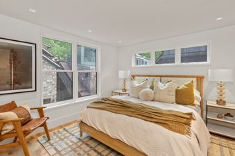 A bright bedroom with a bed adorned with white and gold bedding, flanked by minimalist lamps, with a plaid rug and a leather armchair by the window.