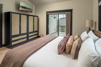 The bedroom at Frosthaven Chalet features a comfortable bed with decorative pillows and a throw blanket, a large wardrobe, a wall-mounted TV, and a glass sliding door leading to a balcony.