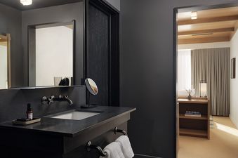 A modern bathroom vanity with a dark countertop and mirror faces an open doorway leading to a sunlit bedroom with light wood accents.