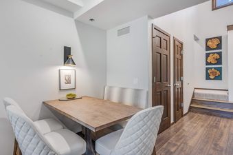 A cozy dining nook with a wooden table, white quilted chairs, and a spotted bench, illuminated by a modern sconce.