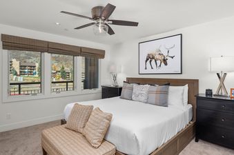 The bedroom at Crestwood 1201 features a king-sized bed, three windows with bamboo shades and mountain views, a ceiling fan, and decorative artwork.