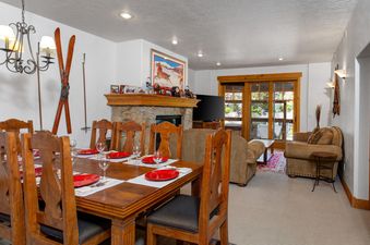 A rustic wooden dining table set with red plates and wine glasses sits in the foreground, with a cozy living area featuring a stone fireplace and plush sofas in the background at Town Pointe C302.