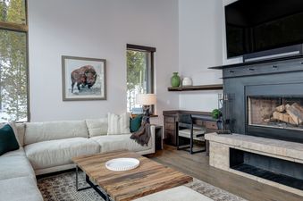 A spacious and elegantly furnished living room at Homestead Chalet 8 Claim Jumper, featuring a light-colored sectional sofa, a wooden coffee table, a fireplace, and large windows looking out onto a snowy pine forest.