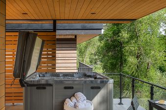 A modern outdoor living space features a hot tub with its cover partially open, inviting guests to relax amidst the lush green trees, creating a serene and private retreat.