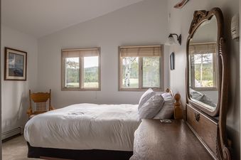 A bright bedroom with a bed, two windows overlooking a natural landscape, and an antique wooden dresser with a mirror.