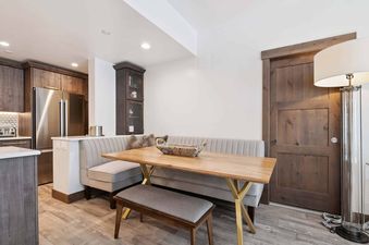 A modern dining area at The Charter D3505 with a wooden table, gold base, and built-in upholstered seating next to a kitchen with wooden cabinets and stainless steel appliances.