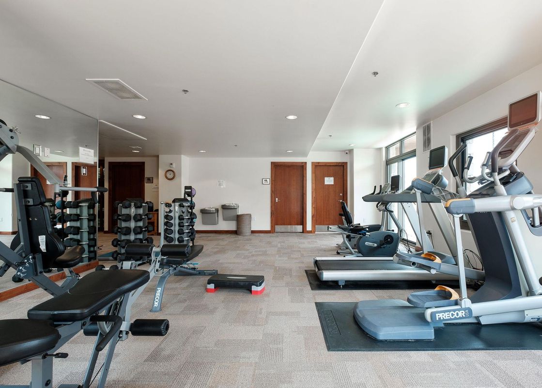 A modern and well-equipped fitness center featuring various exercise machines, including treadmills, ellipticals, a multi-station weight machine, and a rack of dumbbells.