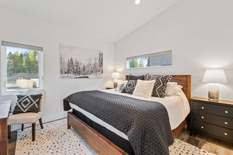 A modern primary bedroom in Quarry B8 features a comfortable bed with a gray comforter, stylish nightstands, elegant lamps, and an armchair, all illuminated by natural light from large windows showcasing an outdoor view.