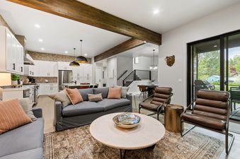 A wide-angle view of the open-concept living room at Dakota Haven, featuring a gray sofa, brown leather chairs, a round coffee table, and a modern kitchen in the background, with a sliding glass door leading to an outdoor patio.
