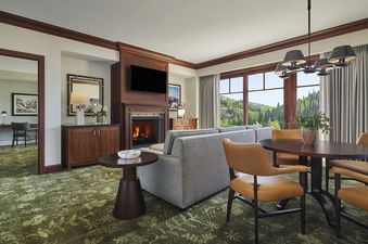 The living area of the Montage Deer Valley One Bedroom Suite Vista View features a comfortable sofa and armchairs, a large flat-screen TV above a lit fireplace, and a dining table with seating for four, all set on a vibrant green botanical carpet.