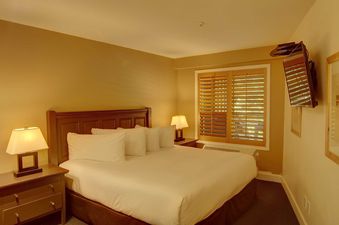 A cozy bedroom with a large bed, two nightstands with lamps, a window with wooden blinds, and a wall-mounted TV.