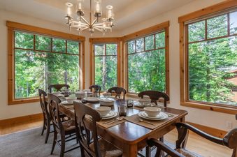 A formal dining room features a large wooden table set for eight with white dishes and placemats, surrounded by classic wooden chairs, all illuminated by a modern chandelier and natural light from expansive windows offering views of lush green trees.