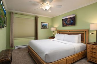 The comfortable king bedroom in Montaneros 204 features a king-sized bed with white linens, wooden furniture, light green walls, a ceiling fan, and artistic landscape prints.