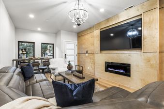 A living room with a large leather sectional sofa, a white armchair, and a coffee table. The room features a fireplace and a large flat screen TV.