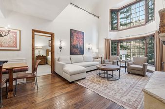 The spacious living area at The Charter G230 is adorned with high vaulted ceilings, a cozy light-colored sectional sofa, two armchairs, a circular coffee table, and large bay windows offering views of snow-dusted trees, complemented by a vibrant abstract