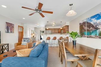 An inviting open concept living area features a blue sofa, a wooden dining table, and a well-equipped kitchen, all bathed in warm lighting.