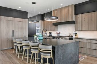 A modern kitchen with a large dark kitchen island, light wood and dark gray cabinetry, and circular pendant lights.