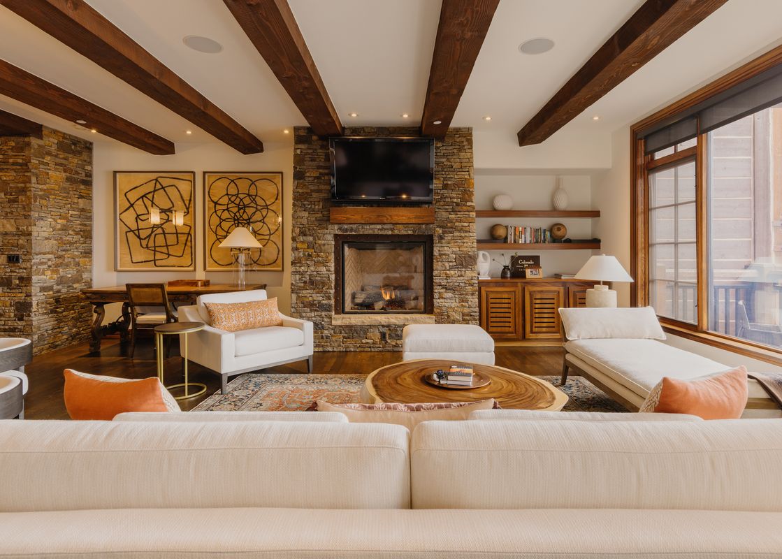 The Peaks Edge at See Forever's elegant living room with a stone fireplace, exposed wooden beams, plush seating, and a large window.