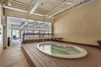 An indoor hot tub with an elevated wooden deck area is situated next to a wall, featuring natural light from large windows in the background.