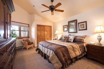 A cozy bedroom at Chateau Chamonix CX142 with an inviting bed, wicker chair, wooden dresser, and framed artworks, creating a warm and rustic ambiance.