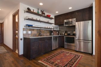 A modern kitchen with dark wood cabinets, stainless steel appliances, open shelving, a stone tile backsplash, and a colorful rug on a wood floor.
