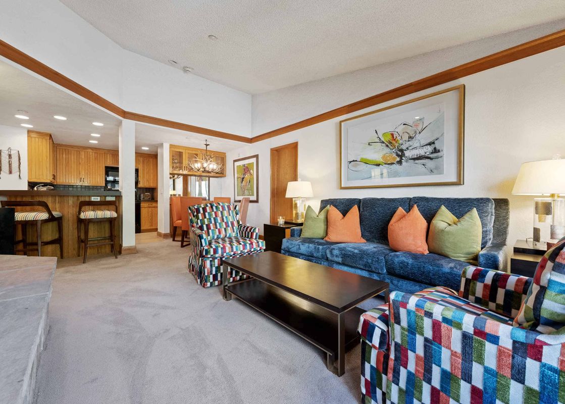 A vibrant living room in The Charter A4505 with a blue sofa and patterned armchairs opens to a dining area with an antler chandelier and an adjacent kitchen.