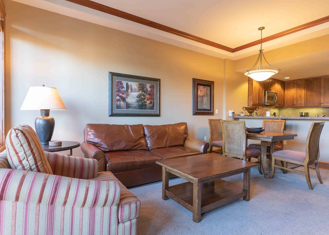 A living room with a large, brown leather couch and a striped armchair, a coffee table, and a dining area in the background.