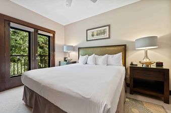 A cozy bedroom at The Charter C2305 features a large bed with white linens, two nightstands with lamps, a nature painting, and a private balcony overlooking lush greenery.