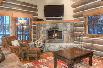 A cozy, rustic living room with a large stone fireplace, comfortable leather furniture, and a view of a snowy forest through the windows.