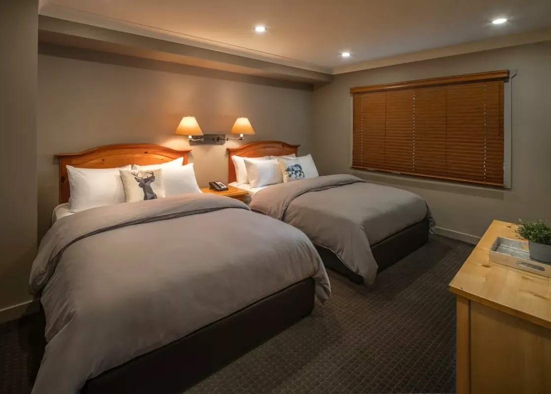 A cozy room at the Buffalo Mountain Lodge Buffalo Suite featuring two queen beds with rustic wooden headboards, soft gray bedding, and charming animal-themed pillows, with warm lighting and wooden blinds.