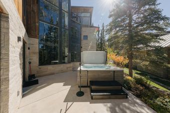 A luxurious outdoor hot tub on the stone patio of Three Peaks, surrounded by pine trees under a bright sun.