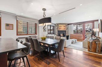 A spacious, open-concept dining room and living area at Solaris 3B West features a large dining table with grey chairs under a modern chandelier, and a living area with a fireplace and windows looking out onto a snowy mountain scene.