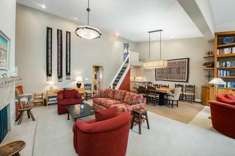 A spacious living area with two red armchairs, a red sofa, a black coffee table, and a fireplace to the left, with an adjacent dining area featuring a wooden table and chairs under a long rectangular light fixture, and a kitchen area beyond a small stairc