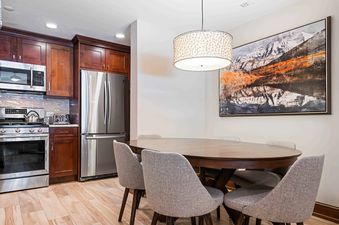An oval wooden dining table with six upholstered chairs sits beneath a modern drum pendant light, adjacent to a kitchen with dark wood cabinets and stainless steel appliances.