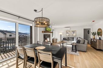 A modern open-concept living and dining area with a dark wood table, comfortable sectional sofa, fireplace, and large sliding doors leading to a balcony with mountain views.