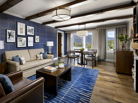 A spacious living area in the Fairmont Chateau 2 Bedroom Valley View Suite features a blue patterned wall, comfortable sofas, a dining table, and a fireplace, with large windows offering a view of the mountains.