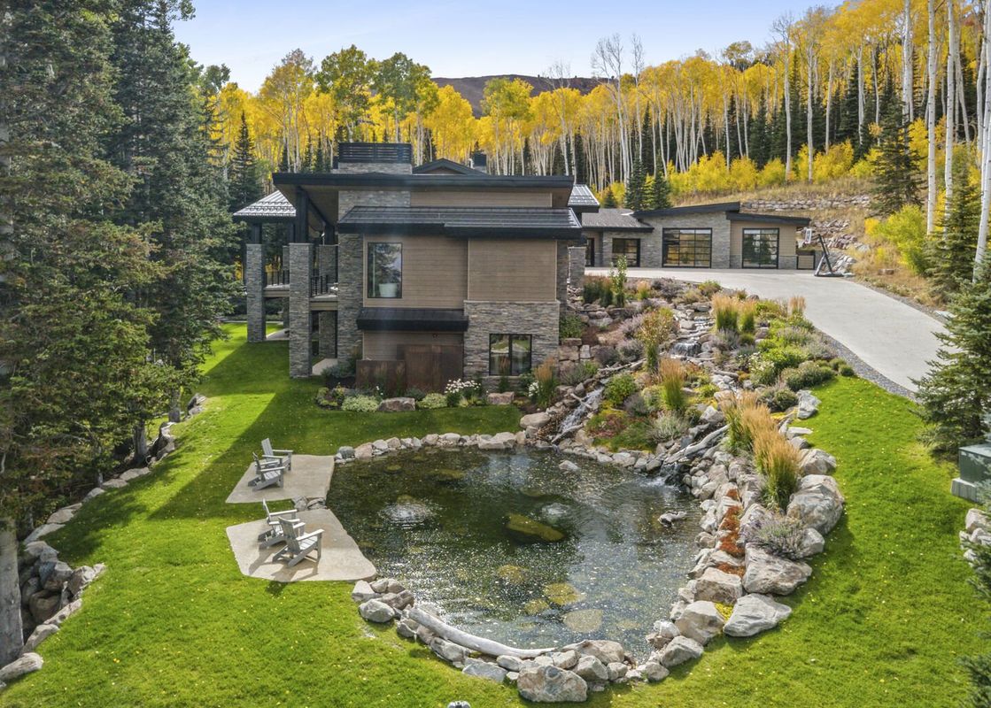 An aerial view showcases the luxurious All In at The Colony mountain estate, featuring contemporary architecture, a tranquil pond with a waterfall, lush landscaping, and a driveway leading to a garage, all set against a backdrop of golden aspen trees and