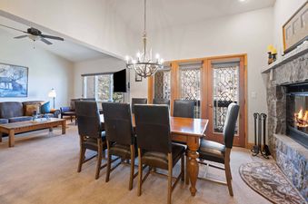 A spacious dining area with a wooden table and leather chairs, illuminated by an antler-style chandelier, adjoins a cozy living area with a stone fireplace and views of the outdoors through ornate windows in The Charter F4405.
