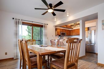 A dining room with a wooden table and chairs, a ceiling fan, and an open doorway leading to a kitchen with wooden cabinets and stainless steel appliances.